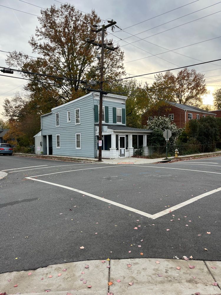 An early 20th century 2 story shotgun style house with pale green siding and dark green shutters. It has an add-on semi-enclosed porch on the front and additional rooms on the back and sits flush to the front & side sidewalks on a corner lot in Alexandria, Virginia. 