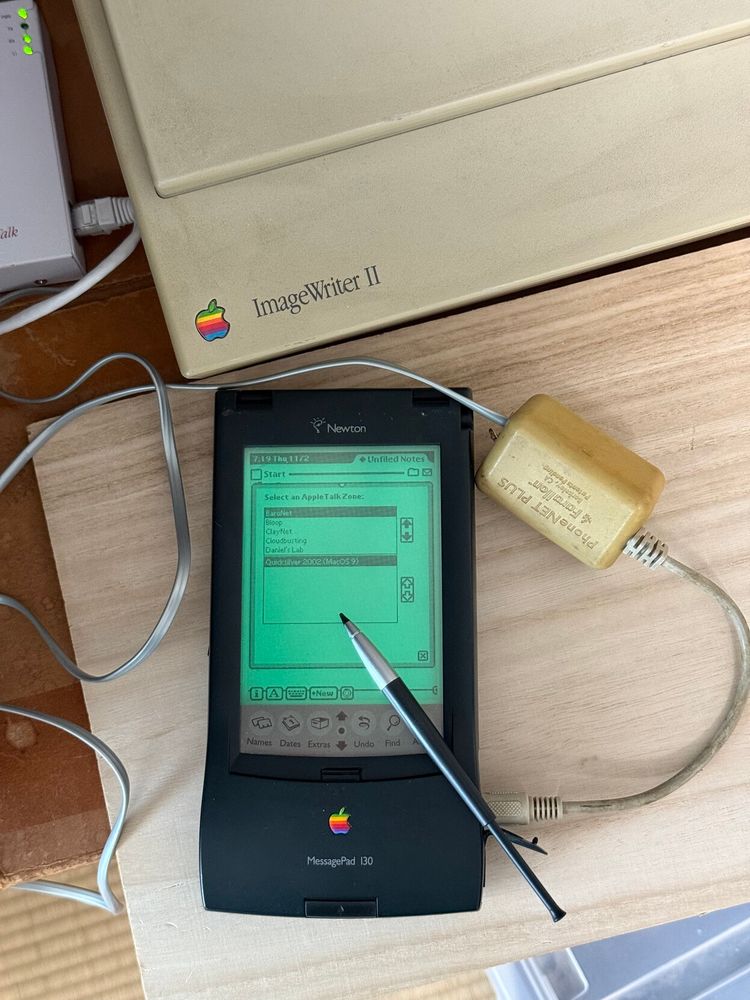 A Newton MessagePad 130 showing a long AppleTalk Zones list, connected to a PhoneNET PLUS adapter. An ImageWriter II and AsanteTalk are also visible in frame