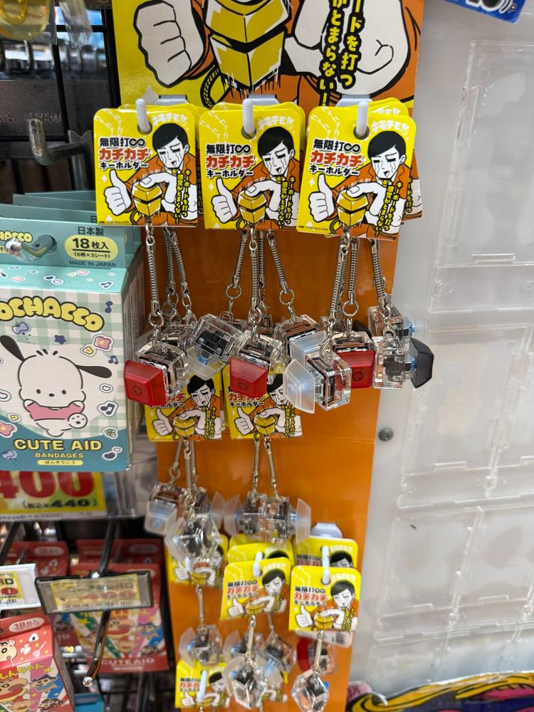 Set of keychains featuring a cartoon character with black hair giving a thumbs up, displayed on a yellow card with Japanese text. The keychain each have a single unmarked keyboard key on them in various colora