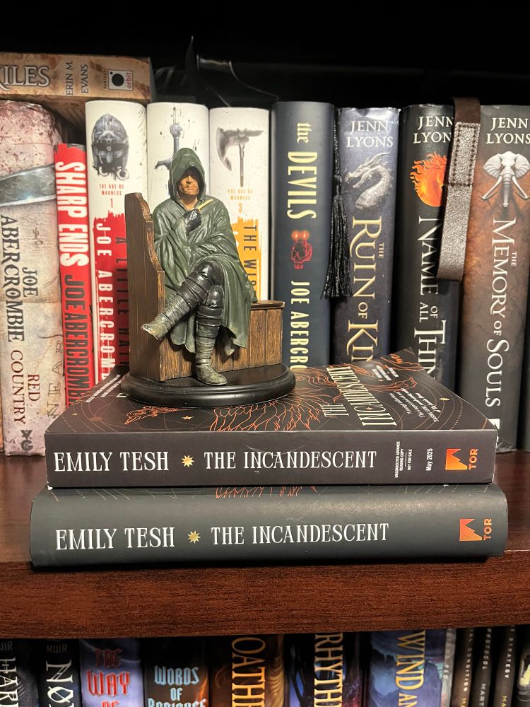 On a bookcase, a statue of Strider sitting upon an ARC copy of The Incandescent and a hardcover copy of The Incandescent by Emily Tesh, various Joe Abercrombie and Jenn Lyons books in the background.
