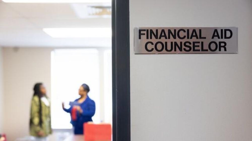 Closeup of a sign reading “financial aid counselor” at the door of a room in which a female staff member speaks to a female student on a college campus.