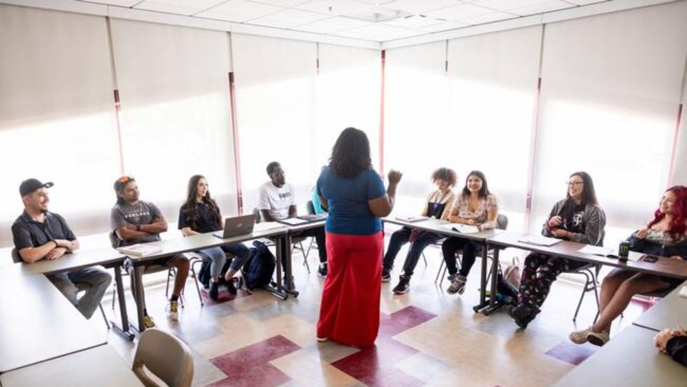 Black female instructor speaking to a multiracial group of students during a class in a sunny classroom on a college campus.