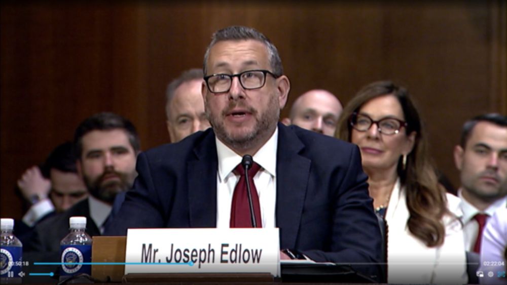 Joseph Edlow, a light-skinned man with gray hair and glasses, wearing a business suit and burgundy tie, speaking at a Senate confirmation hearing. He is sitting at a table and speaking while members of the audience are visible behind him.