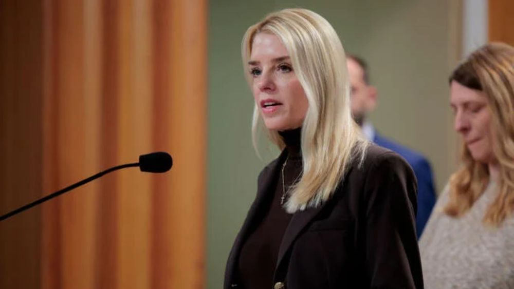 U.S. Attorney General Pamela Bondi stands at a podium and speaks into a microphone. She is a blonde white woman wearing a black suit.