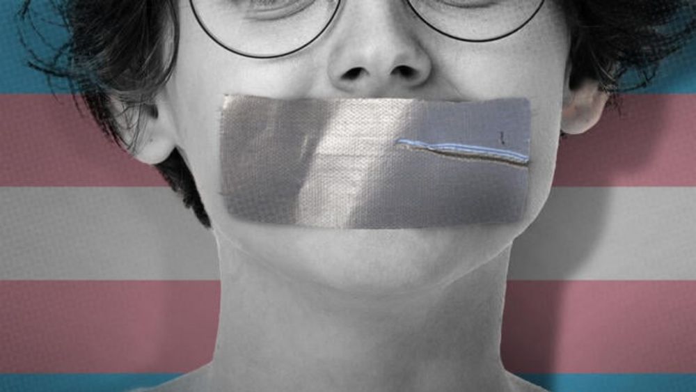 A young person with tape over their mouth in front of a transgender flag.