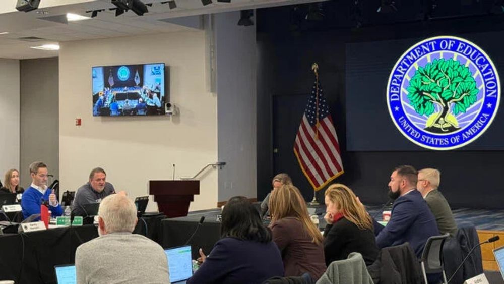 A group of people sit around a table at the Education Department. The agency’s seal is the background.