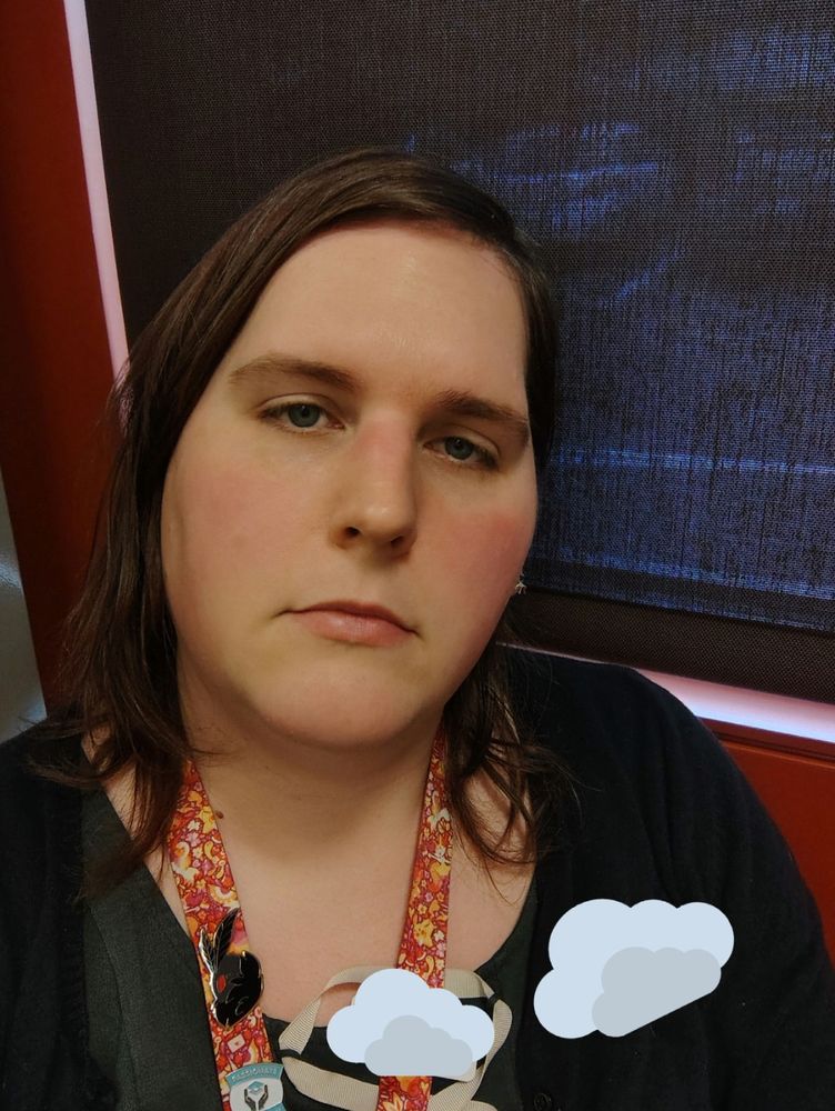 a selfie of me, a transfem non-binary person. I'm wearing a black cardigan, green dress, and a lanyard in the colors of the lesbian pride flag