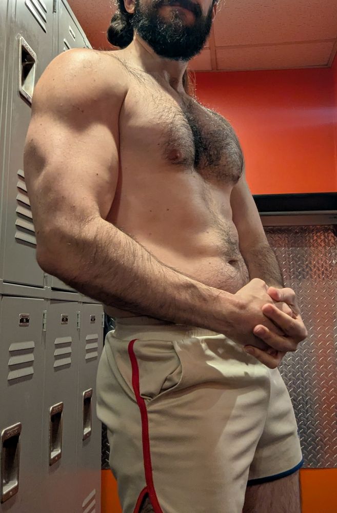 A picture of me flexing in the locker room of the gym. I'm turned a bit away from the camera so my arm is closer to the camera, which makes me look a little bigger. I'm shirtless, wearing my beige raskol shorts with red and blue piping. 