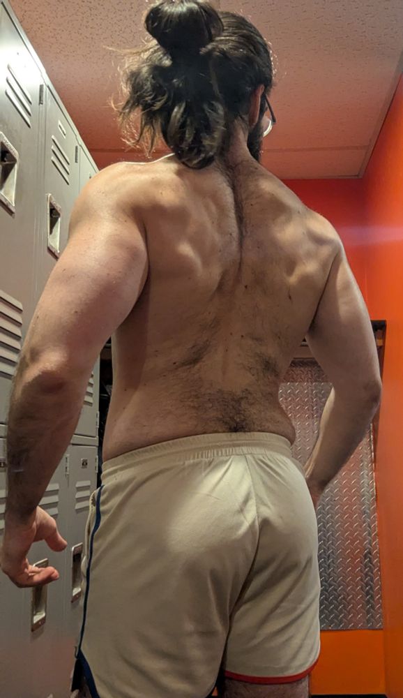 A picture of me flexing in the locker room of the gym. I'm turned away from the camera so you can see my back. I'm shirtless, wearing my beige raskol shorts with red and blue piping. 