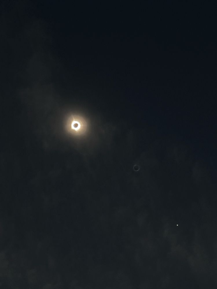 Total solar eclipse a little more zoomed in and darkened. Planet visible to the right. Faint ring of fire lens flare between them.