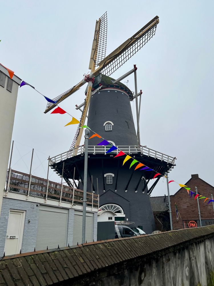 Here you are looking at the windmill in the Arnhem neighbourhood of Klarendaal, de Kroon. It is made of stone, painted grey, and the four blades are not turning at the moment. There is a walkway around the middle section of the windmill and there are bright coloured flags hanging in front of it. The sky is filled with low hanging clouds.