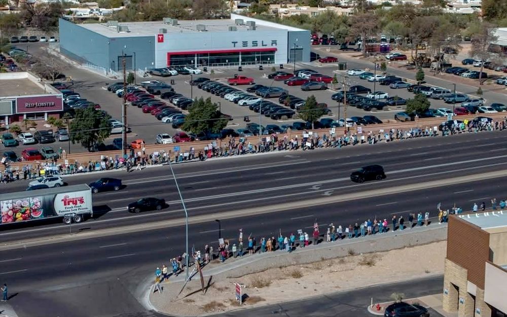 Photo of people lined up with signs on a stroad, in front of and across the stroad from a Tesla dealership. I count eight lanes between protesters on one side and the other