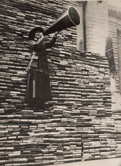 A vintage black and white photo taken during an American Library Association World War One book drive. A librarian in an suit with an ankle-length skirt speaks into a megaphone while standing atop an immense stack of books.