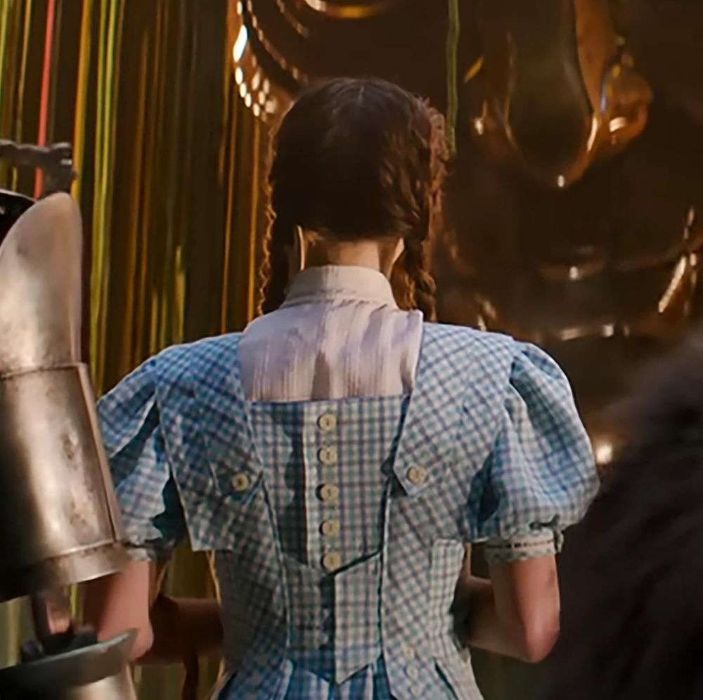 Dorothy known from the wizard of oz in the new wicked movie with her back turned and wearing a blue plaid dress and white undershirt, standing next to the tin man and cowardly lion and they are talking to the great oz