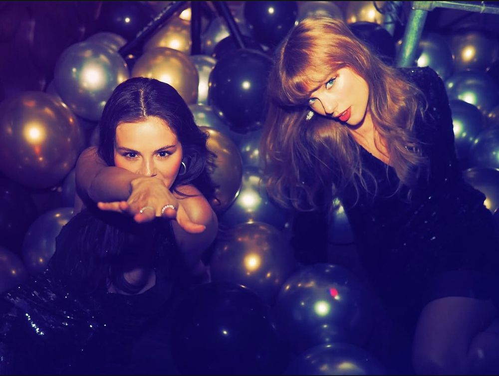 Selena Gomez on the left and Taylor Swift on the right sitting in a pile of balloons that appear to be gold, silver and black. Selena is reaching out towards the camera with rings on her fingers and wearing a black sequin dress and Taylor Swift also appears to be wearing black sequins on her dress as well. Taylor is leaning slightly towards Selena and looking at the camera.