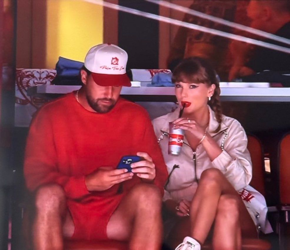 Taylor Swift and Travis Kelce at the Stanley Cup game (hockey). Travis is wearing red and looking at his phone and Taylor is sipping seemingly Diet Coke wearing red lipstick and a white outfit.