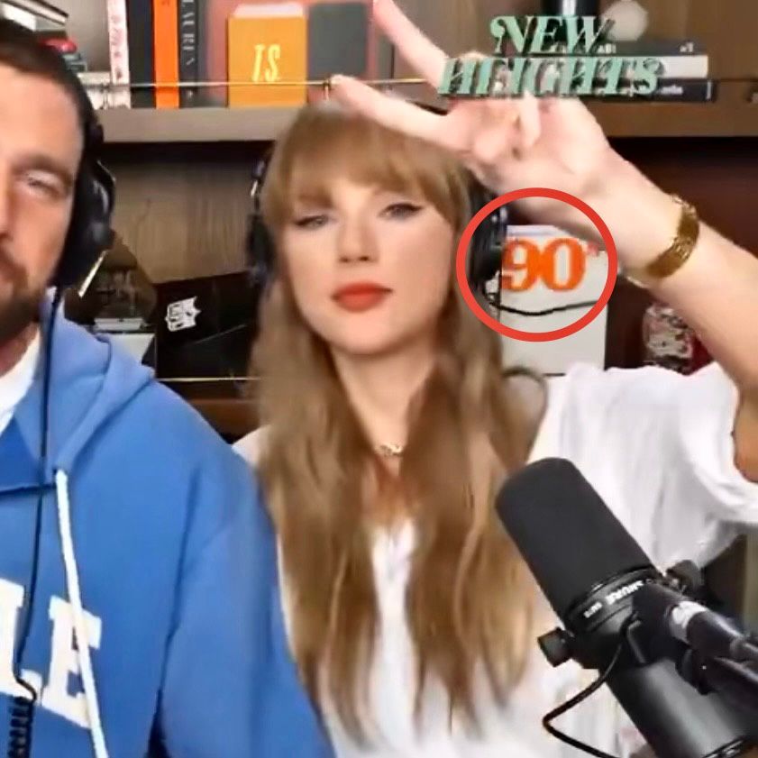 Taylor Swift (and Travis kelce cropped halfway out of frame in blue sweatshirt and headphones) is giving the peace sign in a white shirt and headphones with a mic in front of her revealing a 90s cover or a book or album? in orange font on the shelf behind her. This is a screenshot of the new heights podcast airing tonight!