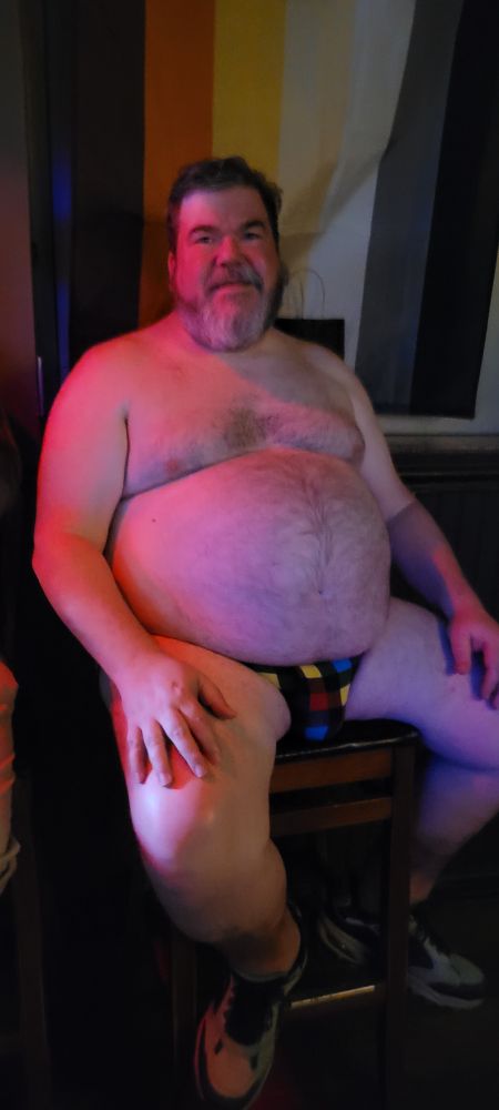 Me on a barstool head-on, lit in red from overhead, in front of a Bear Flag, wearing only a multicolor plaid jockstrap, socks, and sneaks. 