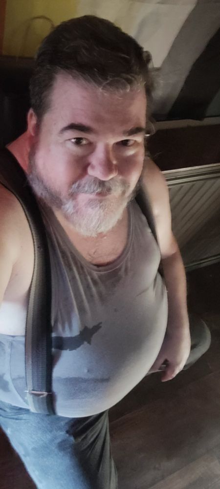 Me on a barstool from overhead, sunlit from a window, in a gray tank top, grayscale striped suspenders, and black jeans. 