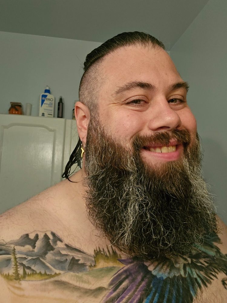 Selfie. Hair is wet and pulled back into ponytail. Sides are shaved. Big brown and grey beard neatly styled. Chest tattoo depicts colorful bird flying over field with trees and mountain in background. Wearing raven wing dangly earrings.