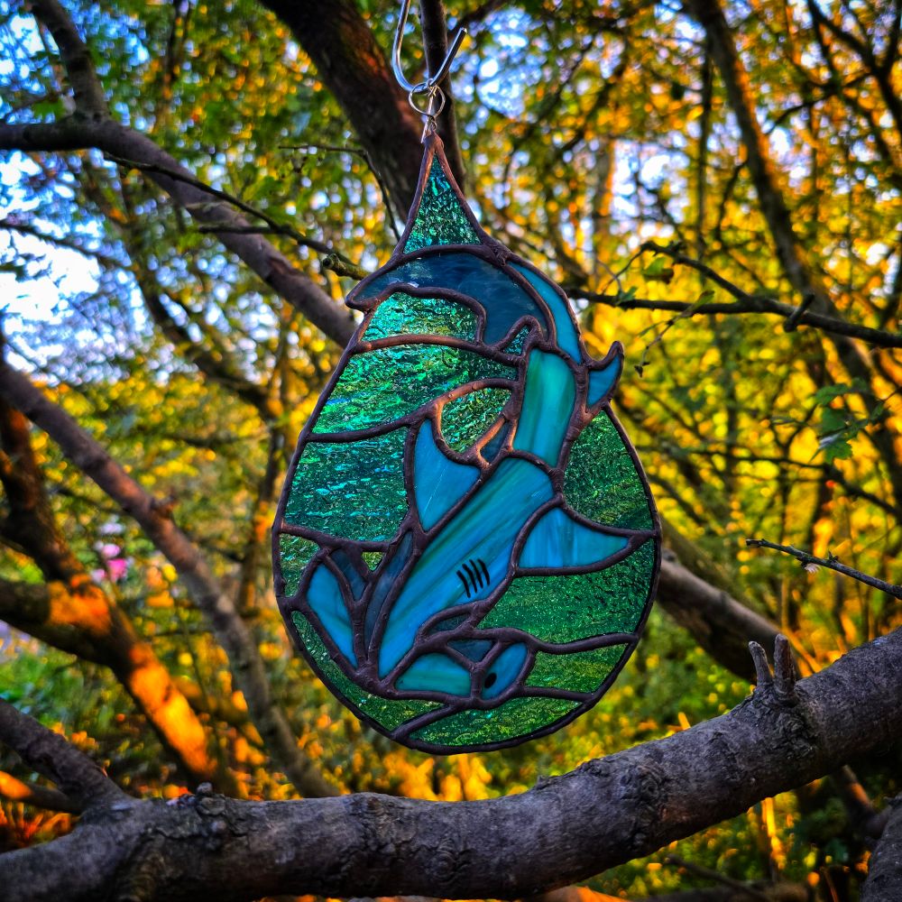 Stained glass ornament shaped like a hammerhead shark, featuring blue and green glass segments, hanging from a tree branch with a backdrop of golden sunlight filtering through dense woodland.