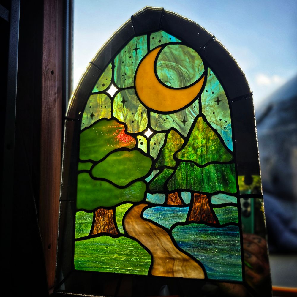 Stained glass panel in an arched shape, depicting a stylized forest scene under a crescent moon. A winding path leads through green trees and across a blue stream, with star-like accents in the sky. Sunlight from behind makes the colors glow, especially the rich greens and golds.
