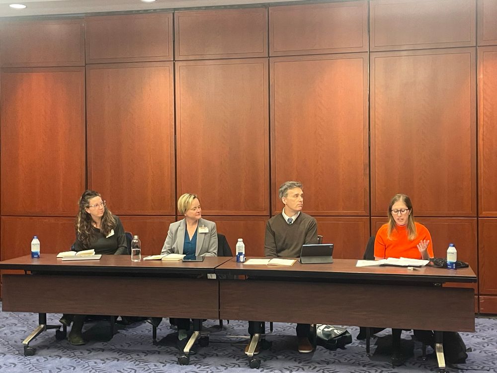 Four speakers at a dais in the front of a conference room. 
