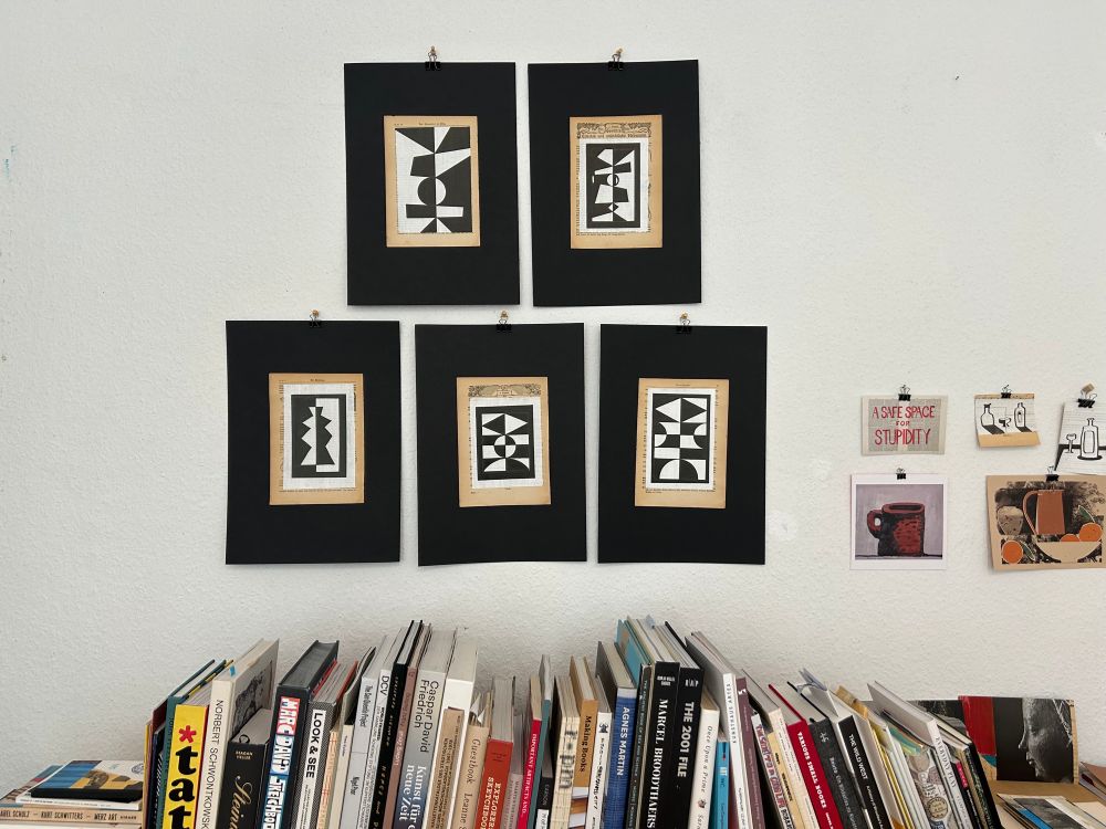 Five black and white found paper collages hanging on a wall above a row of art books. 