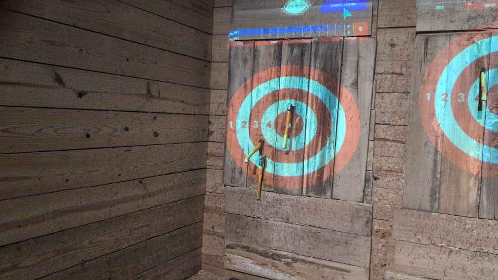 Target on wood wall for ax throwing. 3 axes lodged in wall, one near the bullseye, one on the 3rd ring and the last on the 4th. 