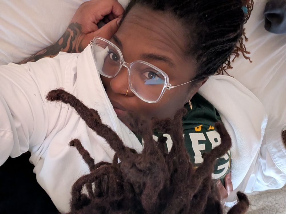 Black woman with locs and a tattoo on her left forearm laying on top of another black woman with clear glasses, sister locs and a Green Bay Packers T shirt. 