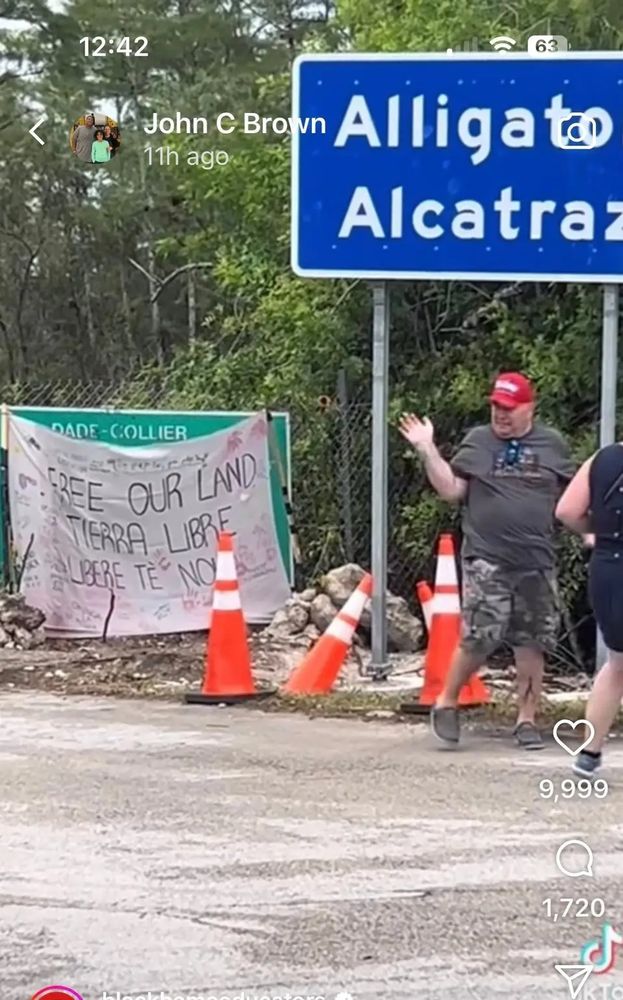 White man in gray shirt and shorts with MAGA hat standing in front of Alligator Alcatraz concentration camp sign 