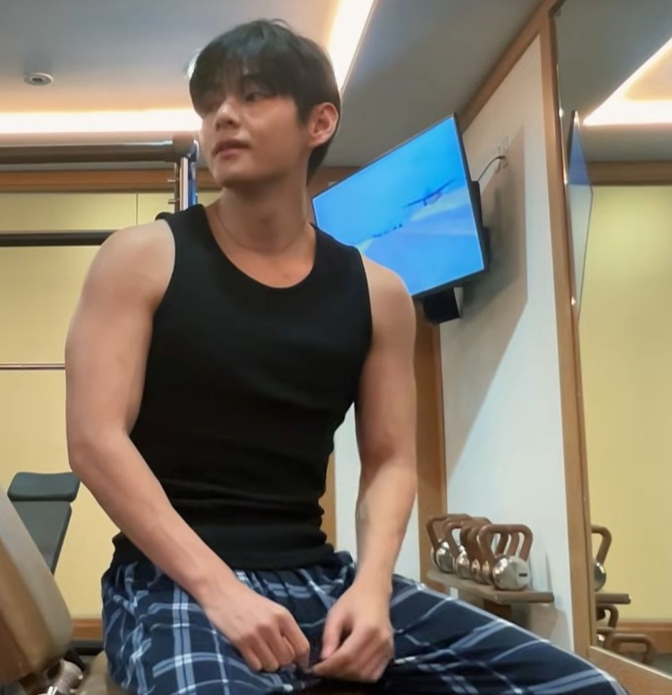 Taehyung in a black tank top at the gym, sitting on a bench with his arms out and looking fine as hell