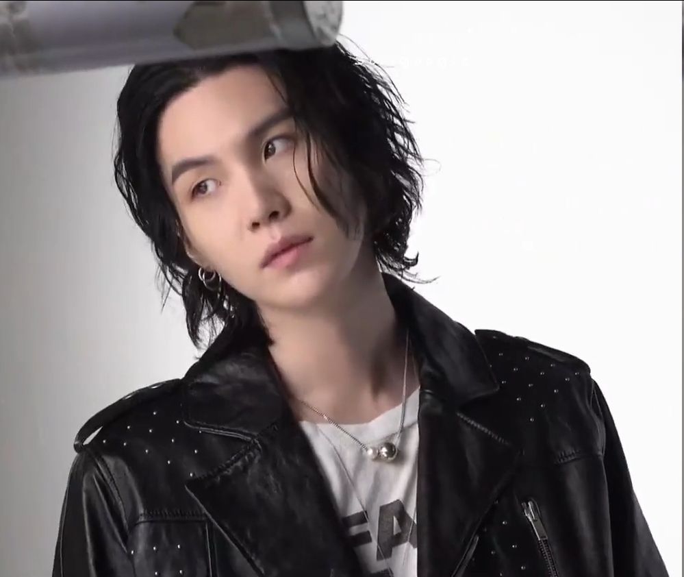 Yoongi against a white background shown from the chest up. He's dressed in the same print t-shirt and studded leather jacket combo from the first photo, his face is turned toward the camera but his eyes are directed toward left.