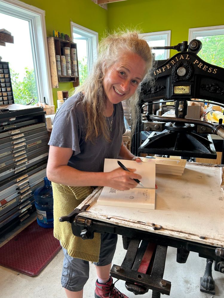 A photo of Monica, a white woman with long curly graying hair, signing copies of her book in the printshop