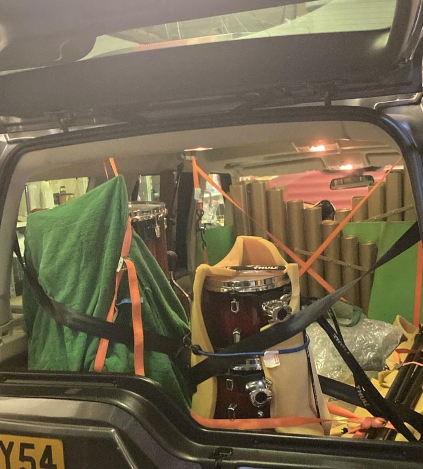 The boot space (seats folded down) of a large SUV entirely taken up with stacks of drums and what likes like organ pipes standing up, criss-crossed with tie downs and straps to keep everything secure. 