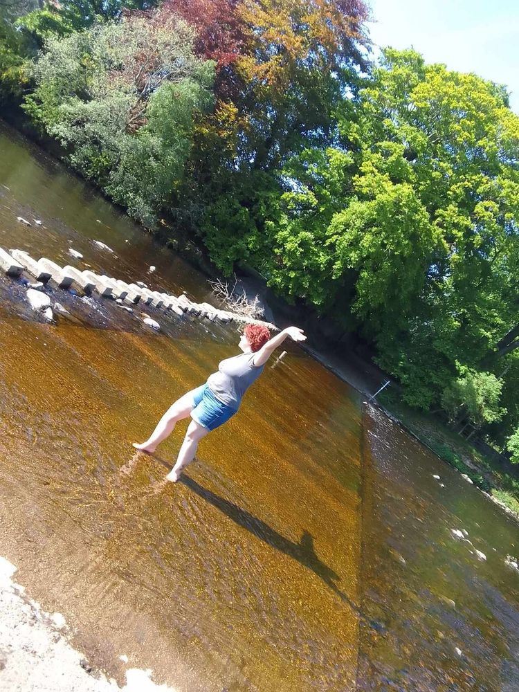 A person with short red hair, grey t shirt, blue denim shorts is dancing on a ford in a river, arms stretched up in a V, although they are standing side on so only one arm is visible. They are facing a row of stones that people can use to walk across from one side to the other. The water is so clear, and it's a lovely sunny day, with very green trees in the background