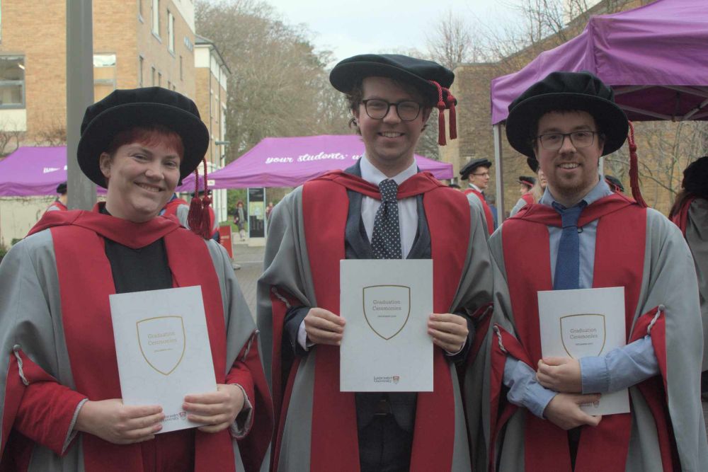 3 PhD graduands, izzy on the left, david in the middle, jamie on the right. They are all wearing silly black cushion hats, and red and grey robes 