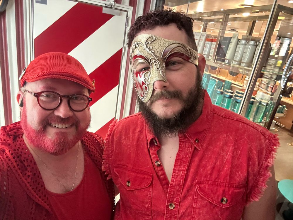 Two men wearing red, one with a red hat and red beard. The other with curly dark brown hair and a mask.