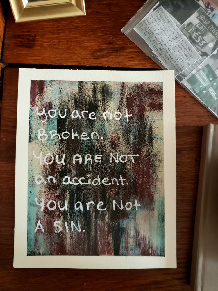 An abstract painting on a wood desk. The painting has brushstrokes in tan, red, black, and teal. Written in white on the painting are the words:
You are not broken.
You are not an accident.
You are not a sin.