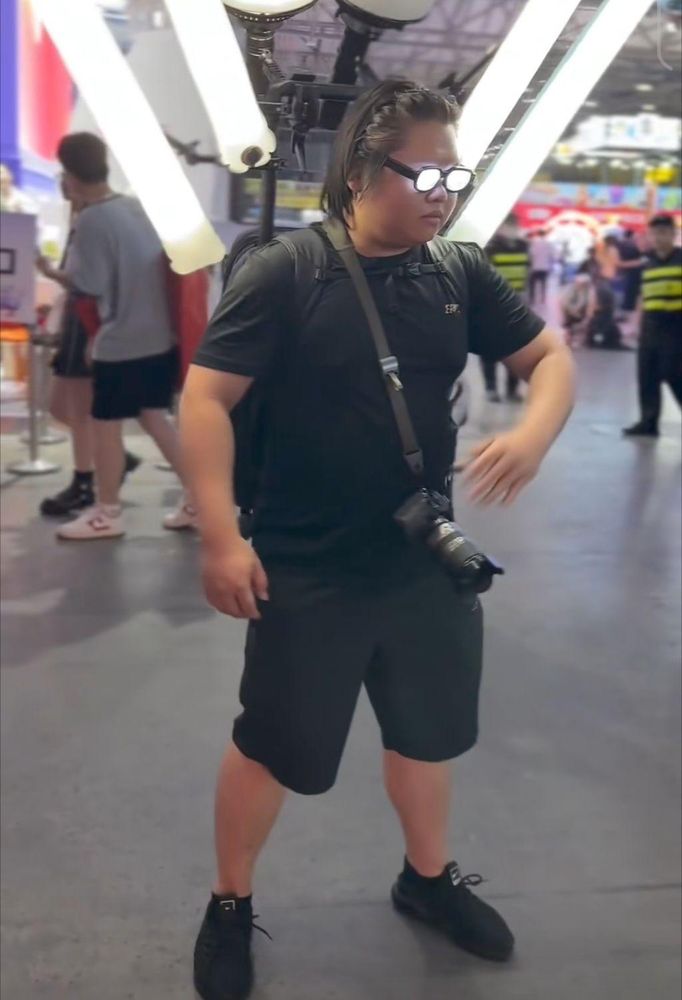 Une personne est au milieu d'un hall, semblant pour une convention ; équipée d'un dispositif supportant plusieurs (trop ?) appareils d'éclairage de photographie ; ainsi qu'un appareil photo professionnel en bandoulière. Les verres de ses lunettes sont éclairées en blanc depuis le coté intérieur, lui donnant une intensité, façon anime. Sa main se porte vers sa caméra avec un flou de mouvement rapide