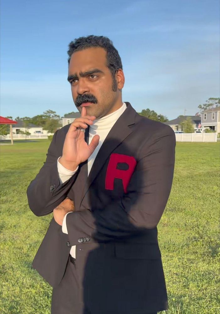 Un homme quadragénaire, est dans une grande étendue d'herbe, quelques maisons au loin. Il arbore une volumineuse moustache et porte une veste de costume-cravate ornée d'un R rouge. Il regarde au loin, un doigt posé sur son menton, l'air pensif. 