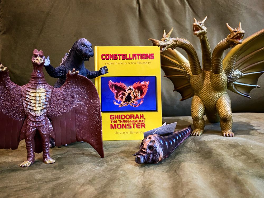On an olive-green sofa, vinyl figurines of Rodan, Godzilla, Mothra (larval form), and King Ghidorah surround a book. The book itself is yellow with red text.