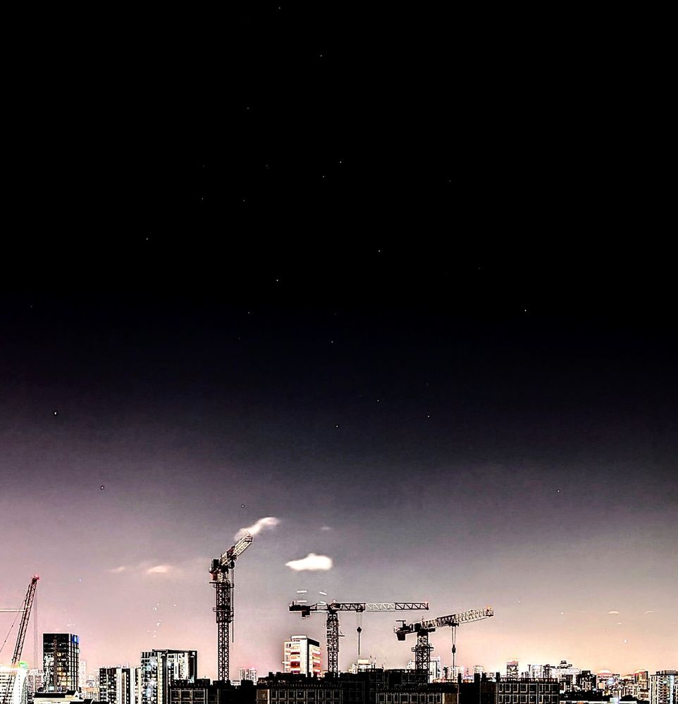 This is a photo of the night sky. The contrast has been heavily adjusted such that the stars can still be visible as white spots in a black night sky despite the light pollution from the city below. We can see a few cranes at a construction site at the bottom of the photo 