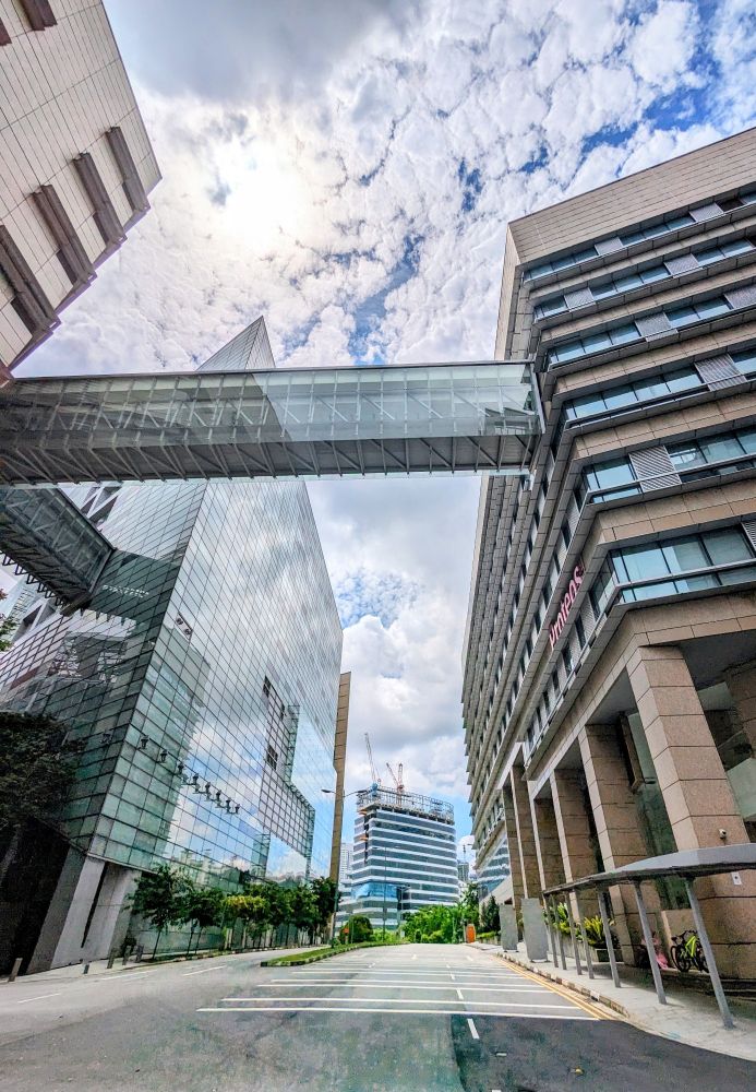 This is a photo of urban landscape with buildings and a skybridge connecting some of the buildings. There is a cloudy sky with patches of blue in the background. The sun is very bright at the top left of the photo while it is behind the white clouds.