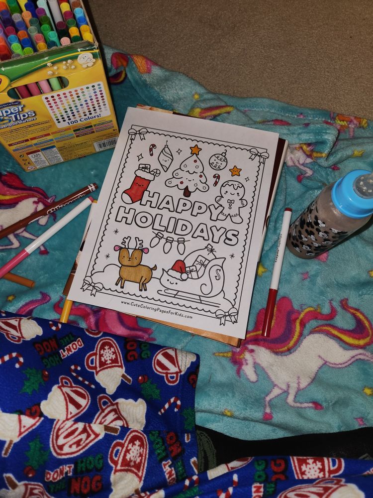 A Christmas coloring page surrounded by markers. A bottle of chocolate milk is nearby. Everything is on a blue unicorn blanket. 