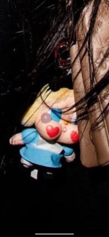 A blonde haired blue eyed doll charm with big cheeks with 2 hearts on them hanging from a brown bag