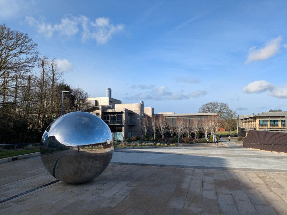 Large metallic ball on a large paved area with building behind and a blue sky.