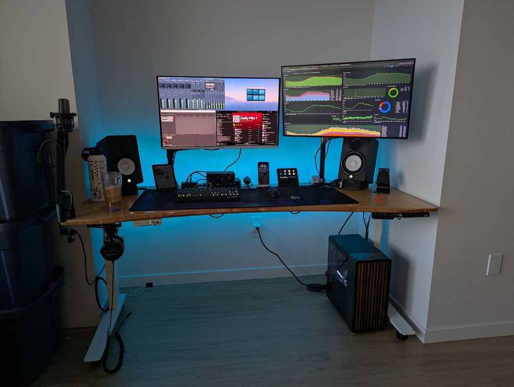 A PC battlestation on a bamboo desk perfectly fit into a nook on a wood vinyl floor with two 32" 4k monitors, a pair of desktop speakers, and a mouse and keyboard. You can see a stack of moving tubs to the left of the desk. A freshly poured glass of monster sits on the left side of the desk.

Blue light washes against the wall behind the desk from a hidden RGB LED strip