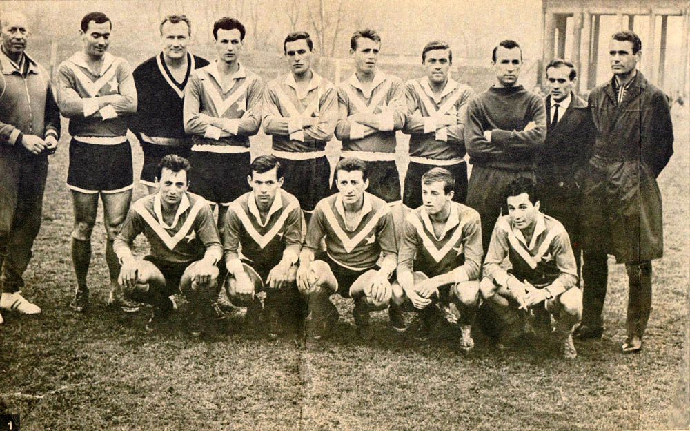 Black-and-white photo of a vintage football team Wisła Kraków taken in 1965. Most men wear matching jerseys with "V" patterns, some standing, others crouching, with two men in suits on the right. The mood is proud and formal.