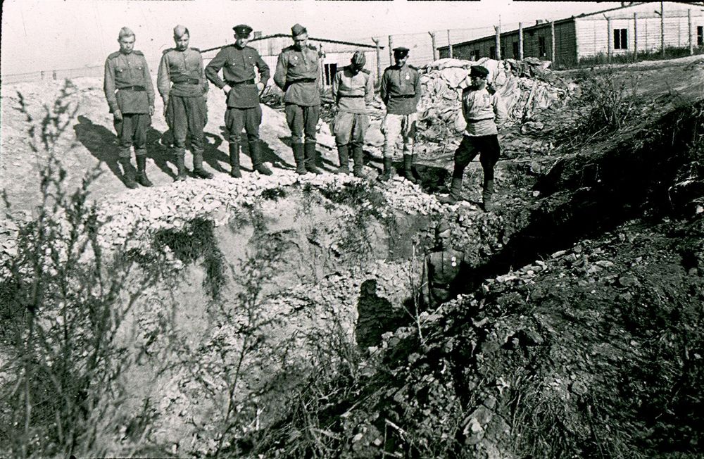 A group of seven soldiers in uniform stand solemnly at the edge of a trench in a barren landscape. Wooden buildings with wire fences line the background.